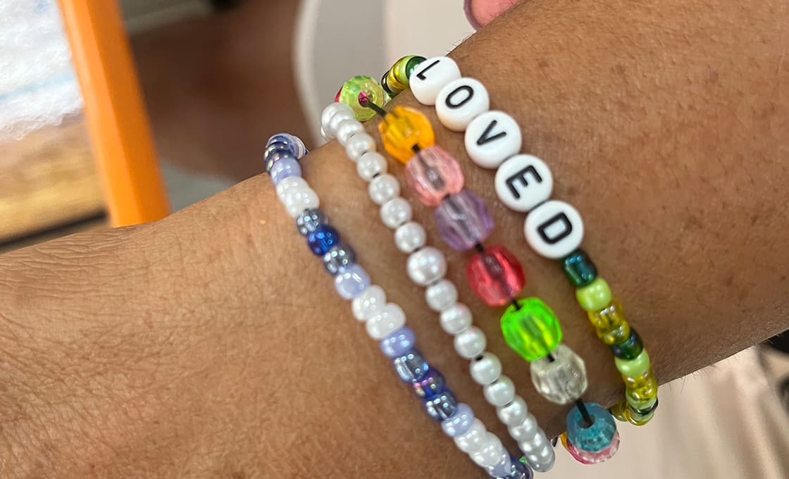 Vier bunte Perlenarmbänder an einem Handgelenk, eines mit weißen Perlen mit der Aufschrift "LOVED".
