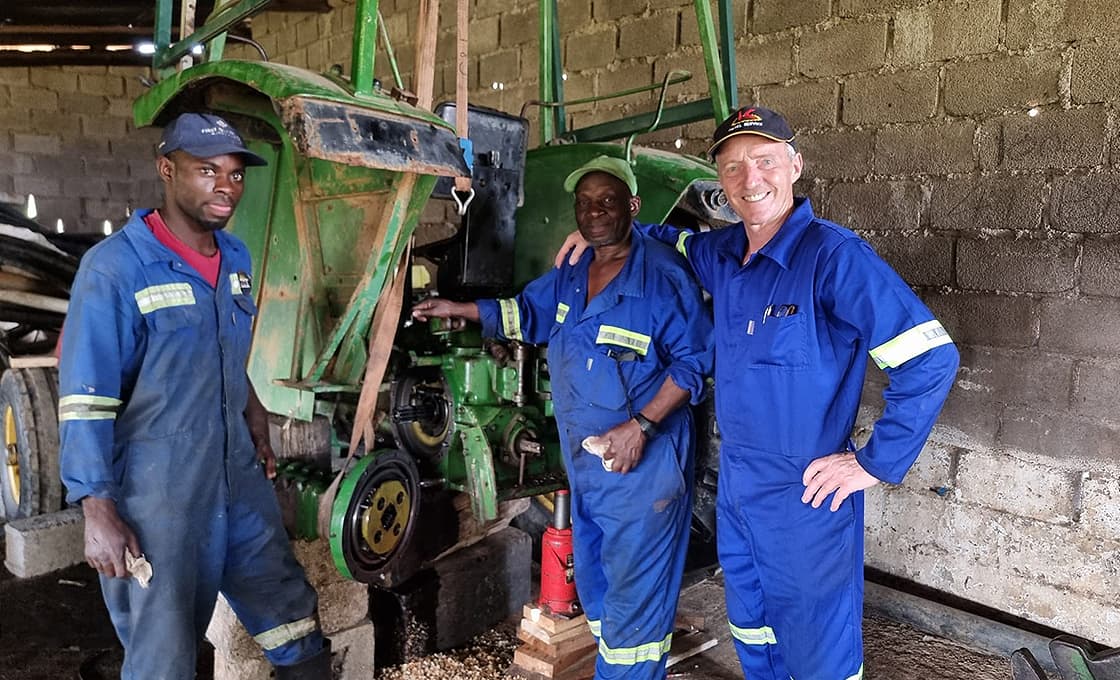Drei Männer in blauen Overalls stehen neben einem teilweise zerlegten Traktor in einer Werkstatt mit Ziegelwänden.