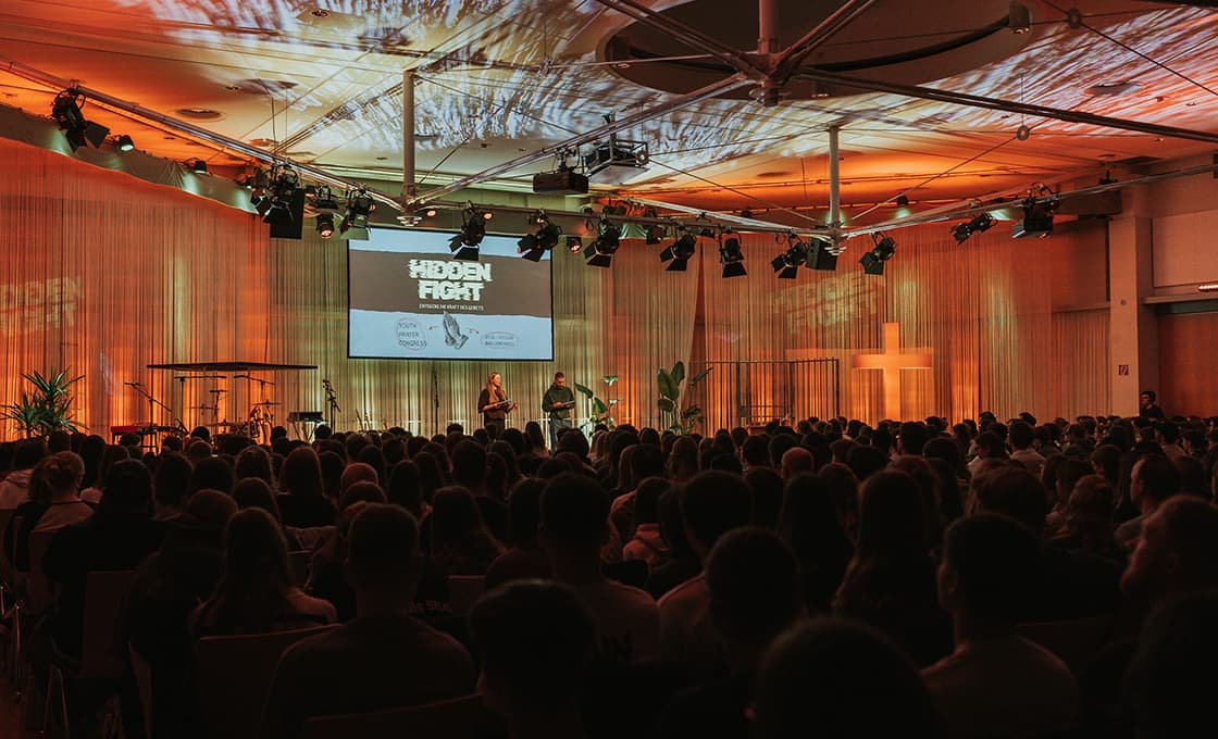 Ein großes Publikum verfolgt eine Bühnenpräsentation mit dem Titel Hidden Fight in einem warm beleuchteten Auditorium mit einem Kreuz an der Wand.