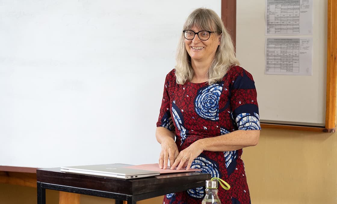 Eine lächelnde Frau mit langen grauen Haaren steht an einem Schreibtisch vor einem Whiteboard, trägt eine Brille und ein rot gemustertes Kleid.
