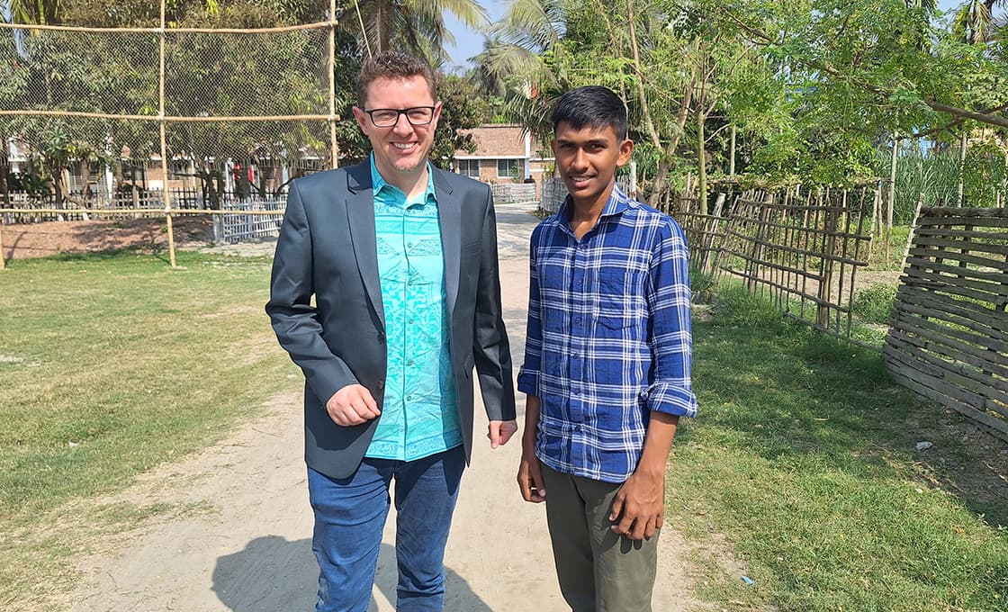Zwei Männer, einer im Blazer, der andere im karierten Hemd, stehen auf einem sonnigen Weg, der von Grünflächen und Zäunen umgeben ist.