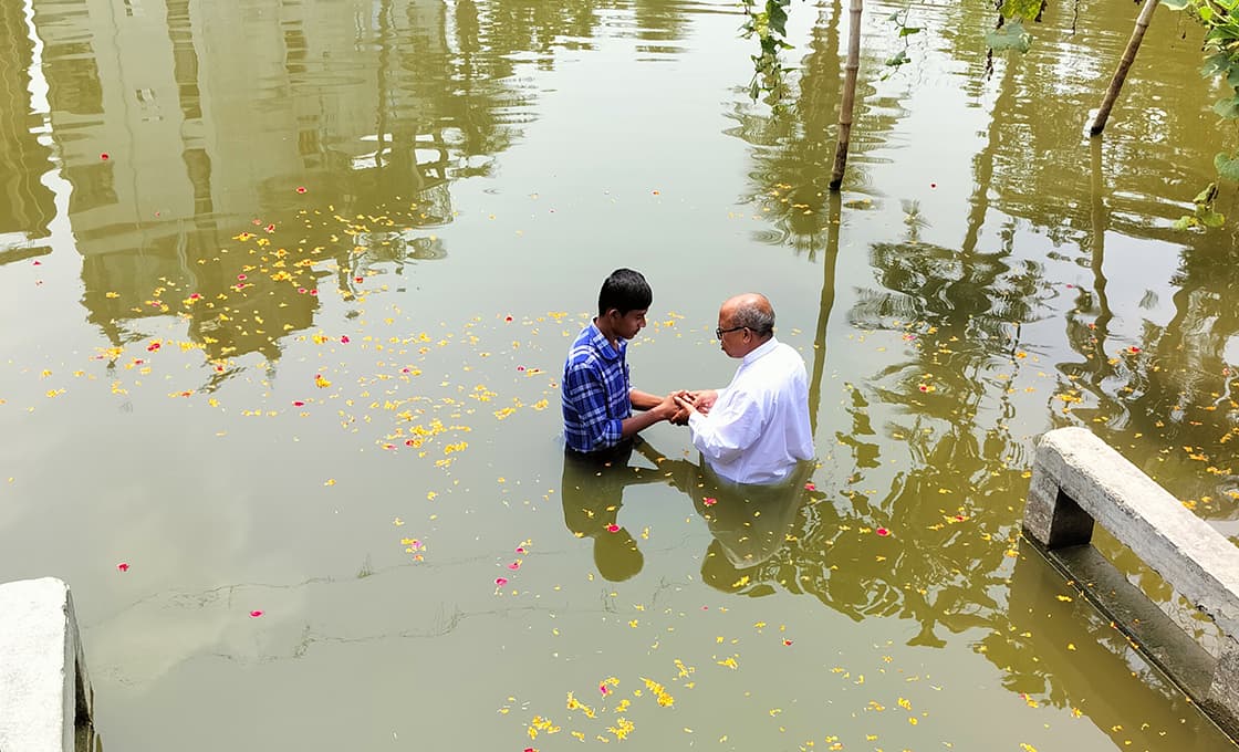 Zwei Männer stehen in hüfthohem Wasser, umgeben von schwimmenden Blumenblättern, und halten sich an einer untergetauchten Bank an den Händen.