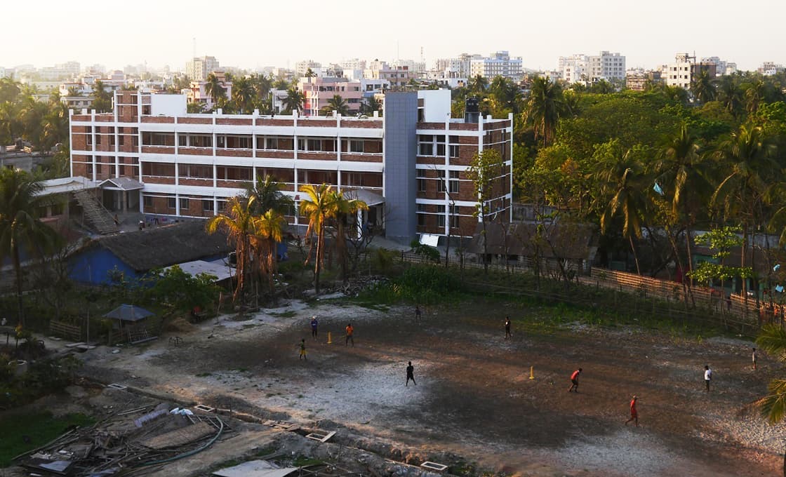Eine Gruppe von Menschen spielt Kricket auf einem unbefestigten Feld in der Nähe eines dreistöckigen Gebäudes und von Palmen in einer städtischen Umgebung.