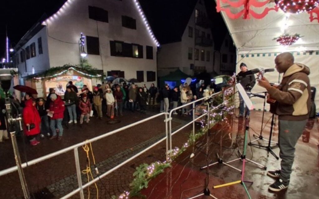 Ein Musiker spielt nachts auf einer Freiluftbühne Gitarre und singt, während eine Menschenmenge ihm bei einer festlichen Veranstaltung zuhört.
