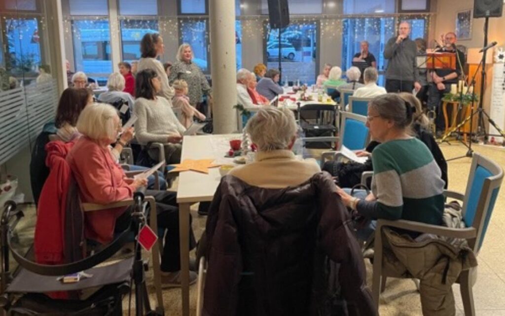 Eine Gruppe von Senioren und Mitarbeitern sitzt an Tischen in einem Gemeinschaftsraum und lauscht den Musikern, die vorne auftreten.